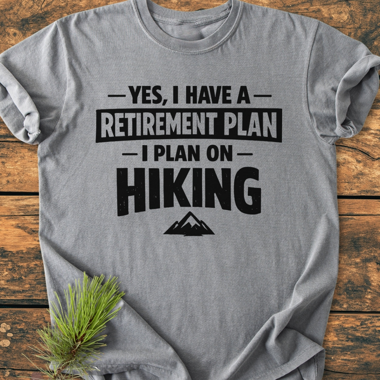 Heather-gray Retirement Plan Hiking - Vintage Wash Tee with bold black text, small mountain graphic, laid flat on wood. Features short sleeves and pine needle accent beside it.