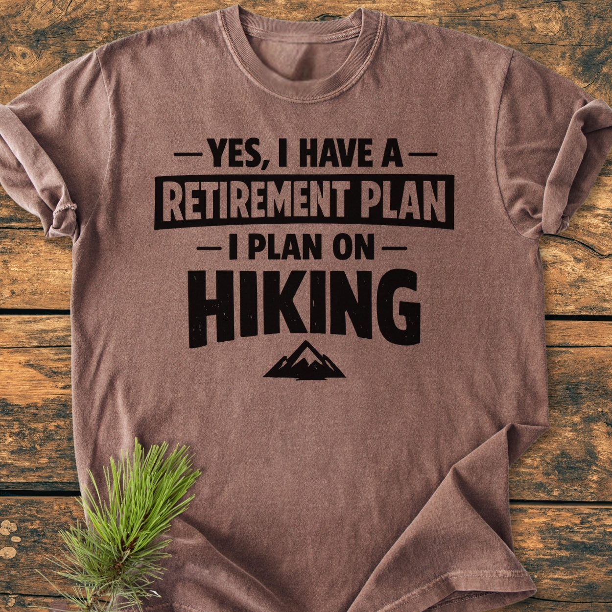 Retirement Plan Hiking - Vintage Wash Tee features bold black text on brown-heather fabric with a mountain icon, showcasing comfort and durability on a rustic wood backdrop with a pine sprig.