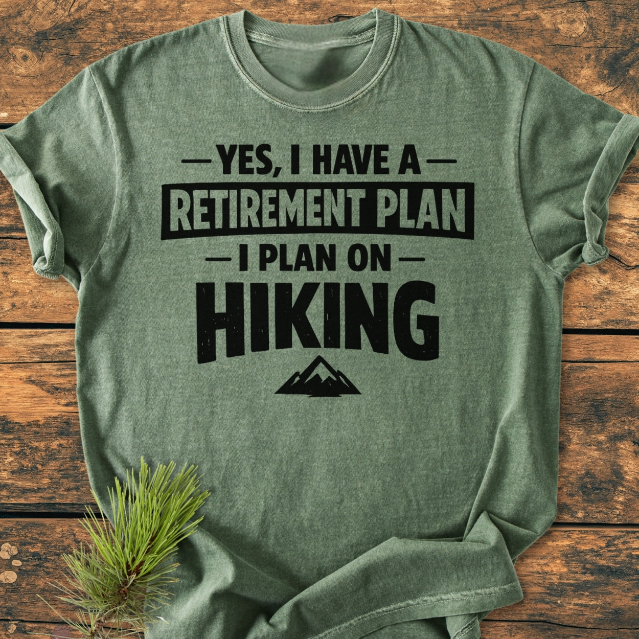 Retirement Plan Hiking - Vintage Wash Tee displayed on wood, features bold hiking-themed text with mountain graphic. A pine sprig accents the soft, heather-green fabric, showcasing its relaxed, durable design.