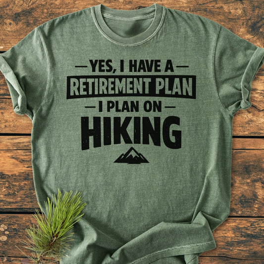 Retirement Plan Hiking - Vintage Wash Tee displayed on wood, features bold hiking-themed text with mountain graphic. A pine sprig accents the soft, heather-green fabric, showcasing its relaxed, durable design.