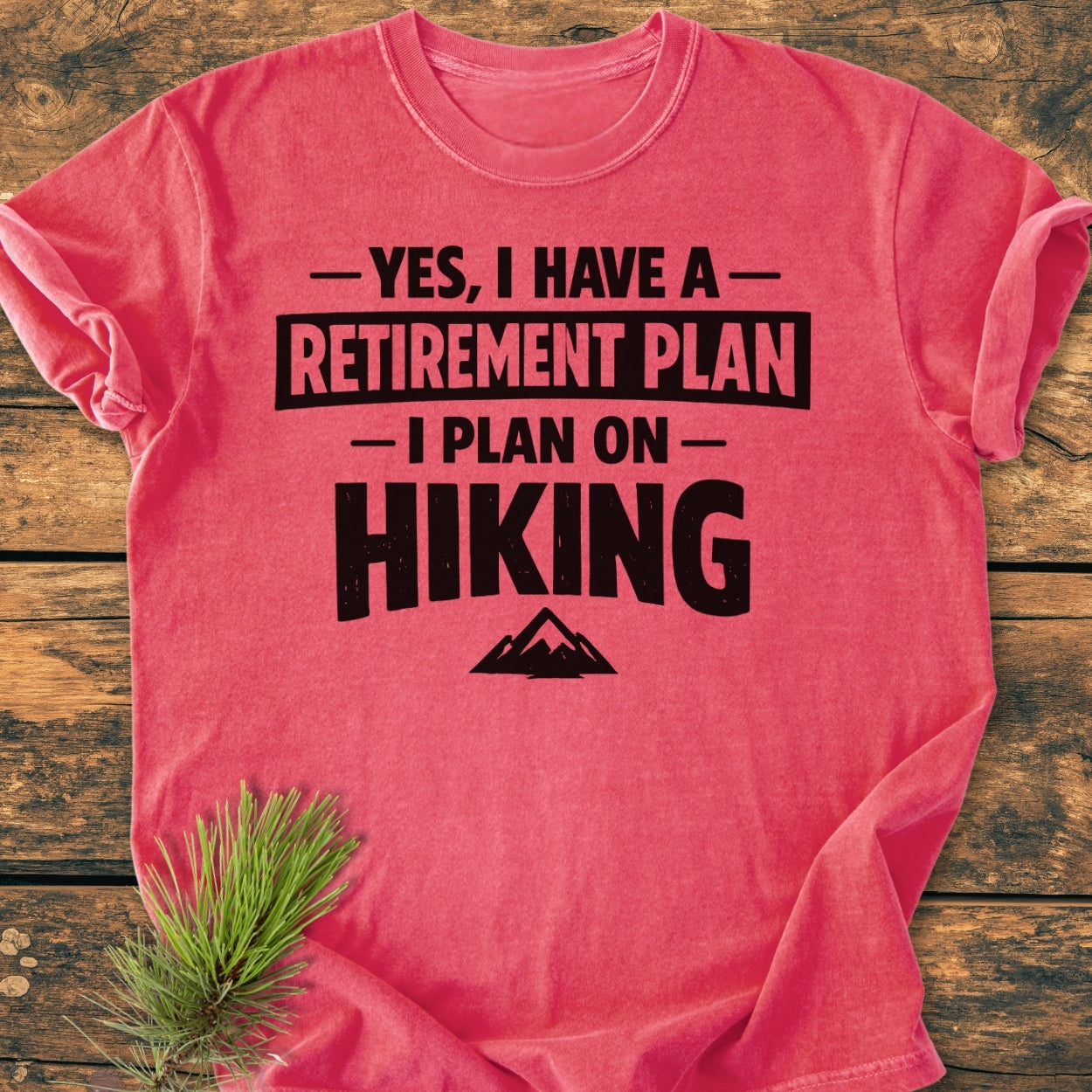 Retirement Plan Hiking - Vintage Wash Tee in coral-red with bold black hiking slogan and mountain graphic, laid on wood. Pine sprig accents corner. Comfortable, durable tee with relaxed fit.