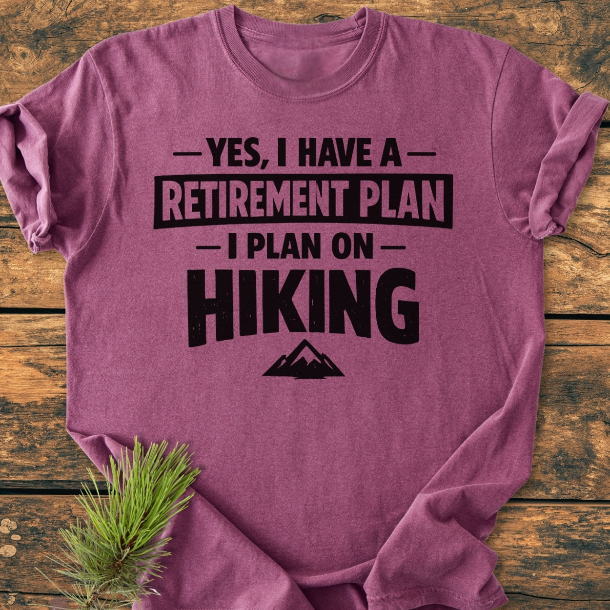 Retirement Plan Hiking - Vintage Wash Tee features a bold print on a maroon tee with a mountain graphic, laid on wood with pine needles, highlighting its relaxed fit and durable stitching.
