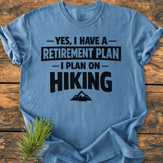 Retirement Plan Hiking - Vintage Wash Tee: Heather-blue, short-sleeve T-shirt with bold lettering and mountain graphic, laid on wood. Soft-washed, 100% ring-spun cotton, relaxed fit, durable stitching, ideal for cozy, casual wear.