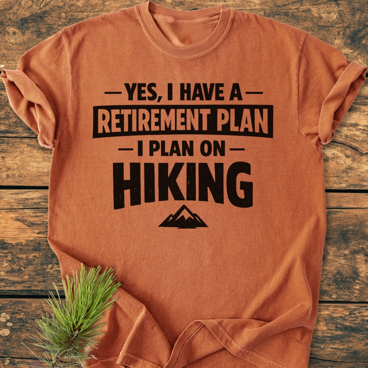 Retirement Plan Hiking - Vintage Wash Tee displayed on wooden boards, featuring bold black text about hiking and a mountain silhouette. A small sprig of pine needles rests nearby.