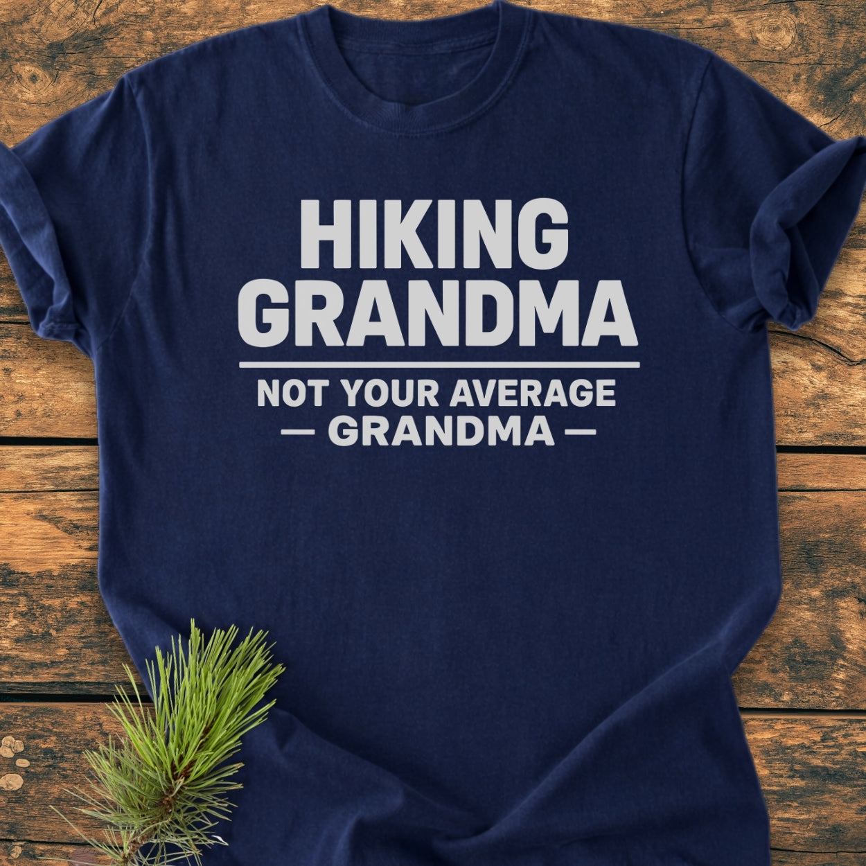 Navy-blue Hiking Grandma - Vintage Wash Tee with bold white text, laid on wood. Features durable, soft-washed 100% ring-spun cotton, relaxed fit, showcasing a pine sprig accent.