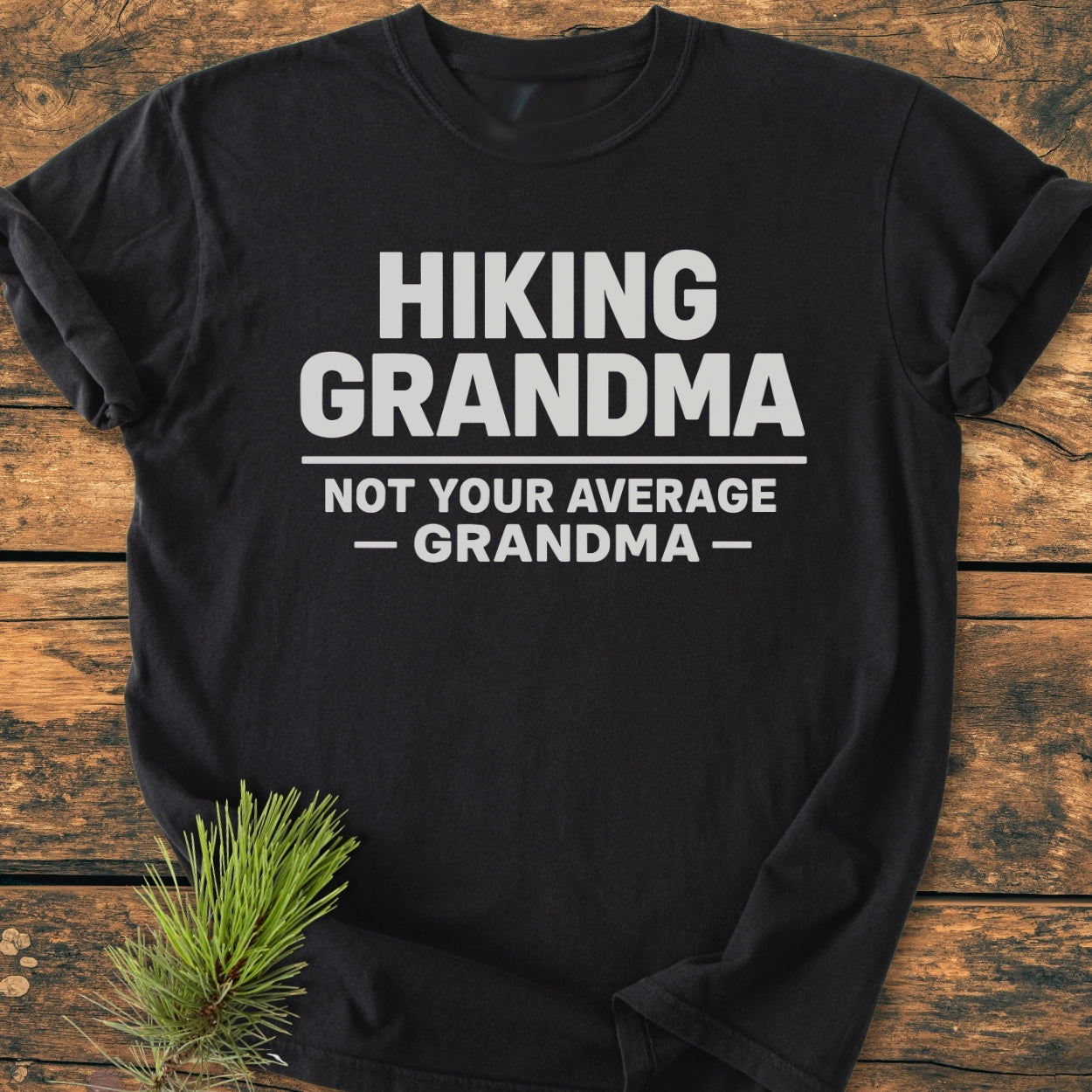 Black T-shirt on wood featuring HIKING GRANDMA — NOT YOUR AVERAGE GRANDMA — in bold white text, beside a pine sprig. The Hiking Grandma - Vintage Wash Tee offers comfort and durability.