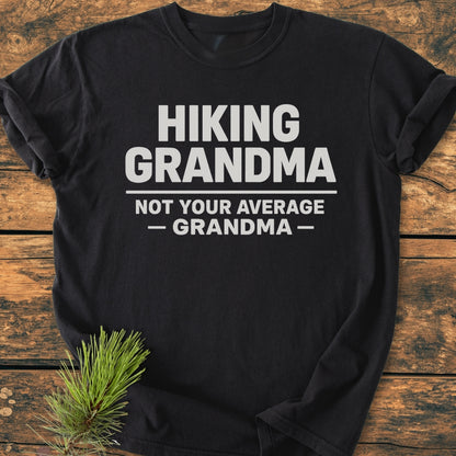 Black T-shirt on wood featuring HIKING GRANDMA — NOT YOUR AVERAGE GRANDMA — in bold white text, beside a pine sprig. The Hiking Grandma - Vintage Wash Tee offers comfort and durability.