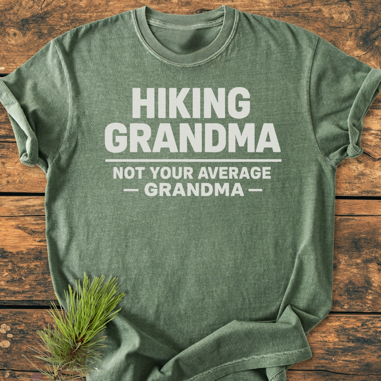 Hiking Grandma - Vintage Wash Tee displayed on wood, featuring bold white text. Accompanied by a pine branch, illustrating the outdoor theme. Made of soft-washed, pre-shrunk, ring-spun cotton for comfort and durability.