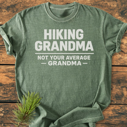Hiking Grandma - Vintage Wash Tee displayed on wood, featuring bold white text. Accompanied by a pine branch, illustrating the outdoor theme. Made of soft-washed, pre-shrunk, ring-spun cotton for comfort and durability.