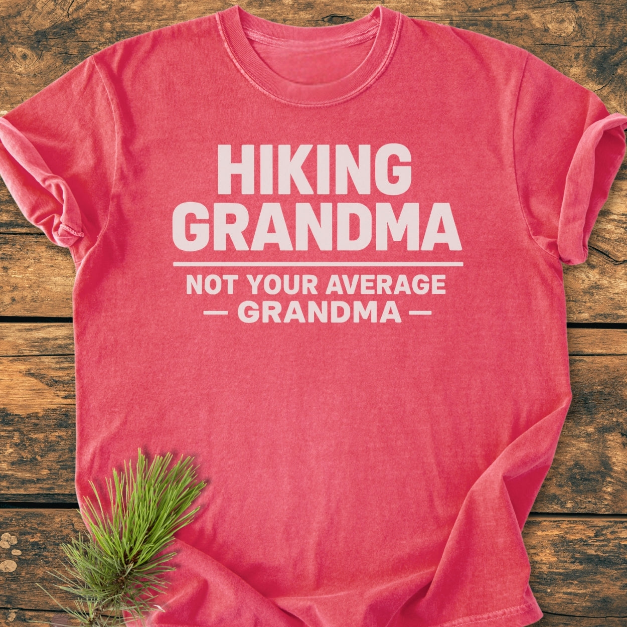 Hiking Grandma - Vintage Wash Tee with bold white slogan on salmon-pink fabric, laid flat. A pine sprig accents the soft, durable design, showcasing the tee's cozy, relaxed fit.