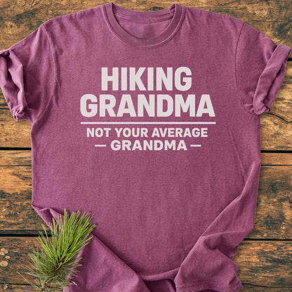 Heather-purple Hiking Grandma - Vintage Wash Tee with bold white text, laid flat on wood. Features soft-washed, garment-dyed cotton for comfort and durability, with a small pine sprig accent.