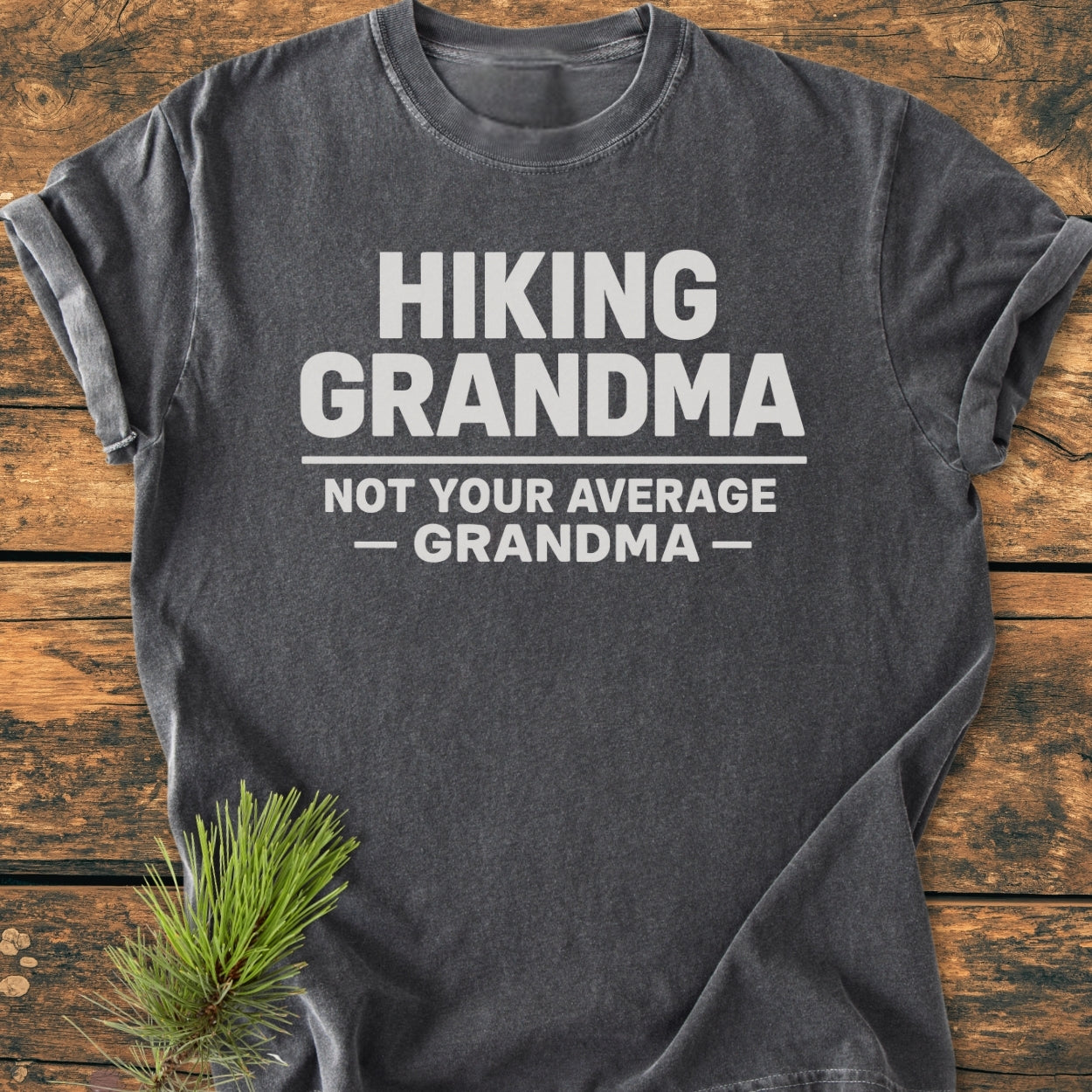 Hiking Grandma - Vintage Wash Tee: Charcoal gray, rolled-sleeve T-shirt with bold white slogan, “HIKING GRANDMA — NOT YOUR AVERAGE GRANDMA —,” laid on a rustic wooden surface with a pine sprig accent.