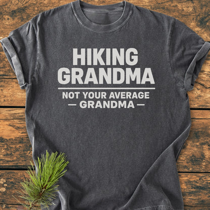 Hiking Grandma - Vintage Wash Tee: Charcoal gray, rolled-sleeve T-shirt with bold white slogan, “HIKING GRANDMA — NOT YOUR AVERAGE GRANDMA —,” laid on a rustic wooden surface with a pine sprig accent.