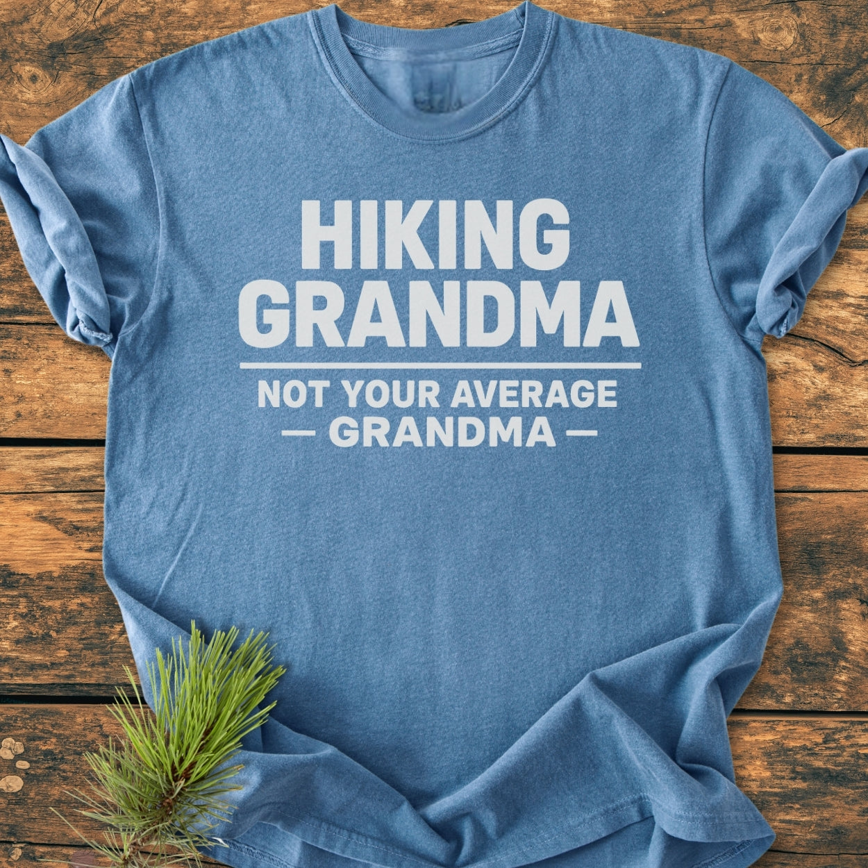 Hiking Grandma - Vintage Wash Tee: A heather-blue T-shirt with bold white slogan Hiking Grandma, Not Your Average — Grandma — printed on front, laid on wood with pine sprig accent.