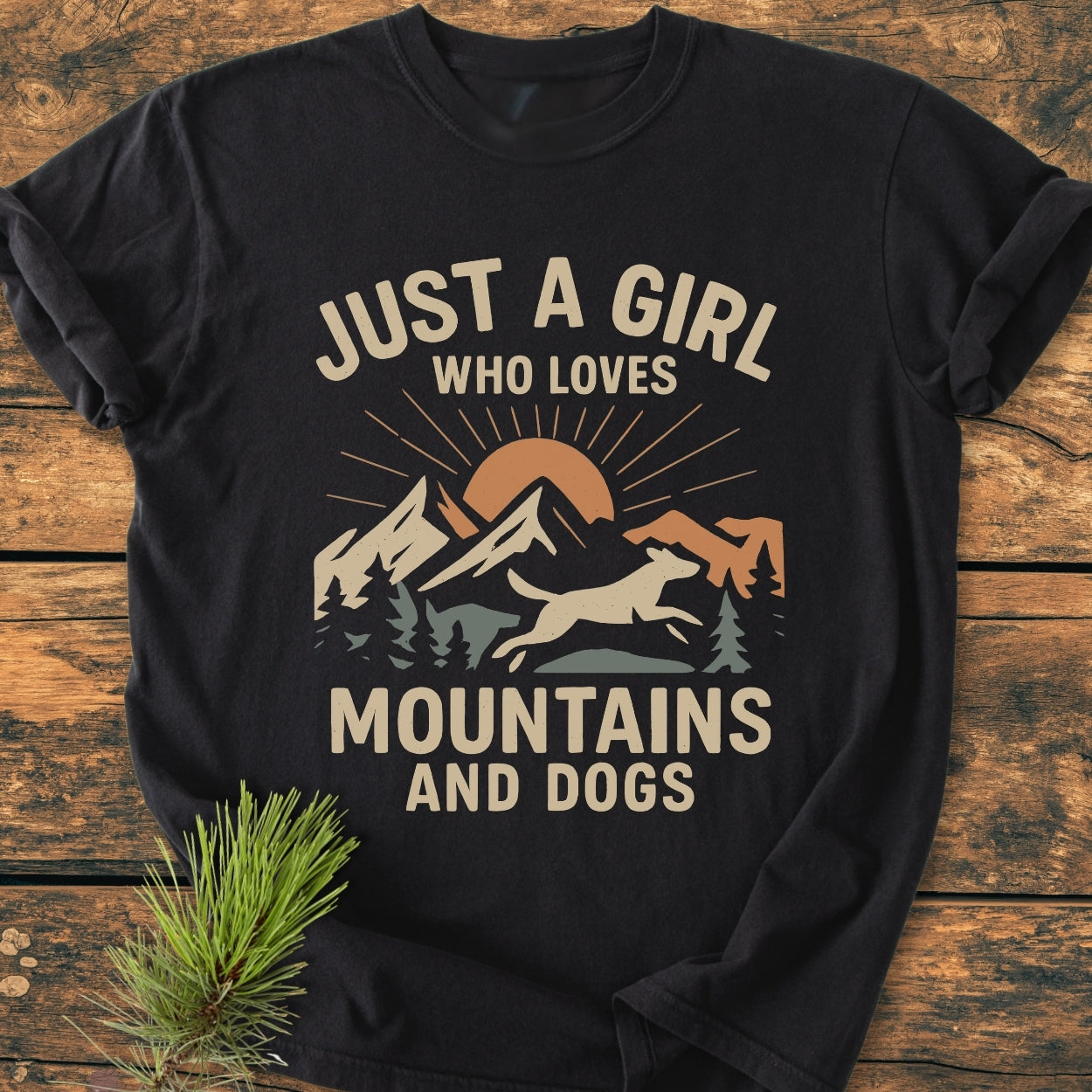 Just A Girl Who Loves Mountains And Dogs - Vintage Wash Tee features a mountain and dog graphic with a slogan, set against a rustic wooden backdrop, accompanied by a small evergreen sprig.