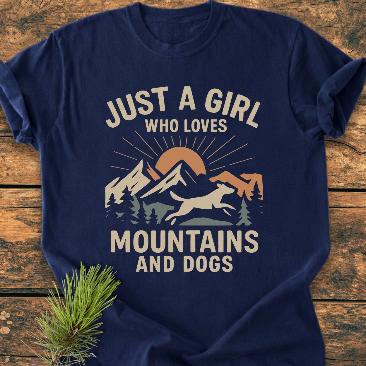 Just A Girl Who Loves Mountains And Dogs - Vintage Wash Tee features a mountain, trees, dog silhouette, and sun graphic on a navy-blue shirt, laid on wood with a pine sprig beside it.