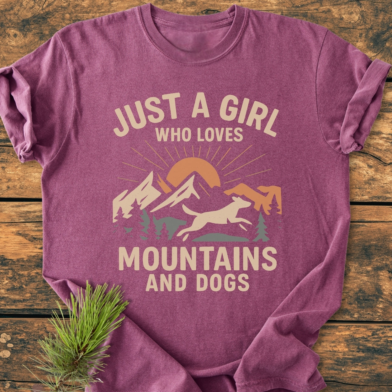 Just A Girl Who Loves Mountains And Dogs - Vintage Wash Tee, folded on wood with cream print of mountains, trees, sun, and running dog graphic, conveying outdoor passion and cozy style.