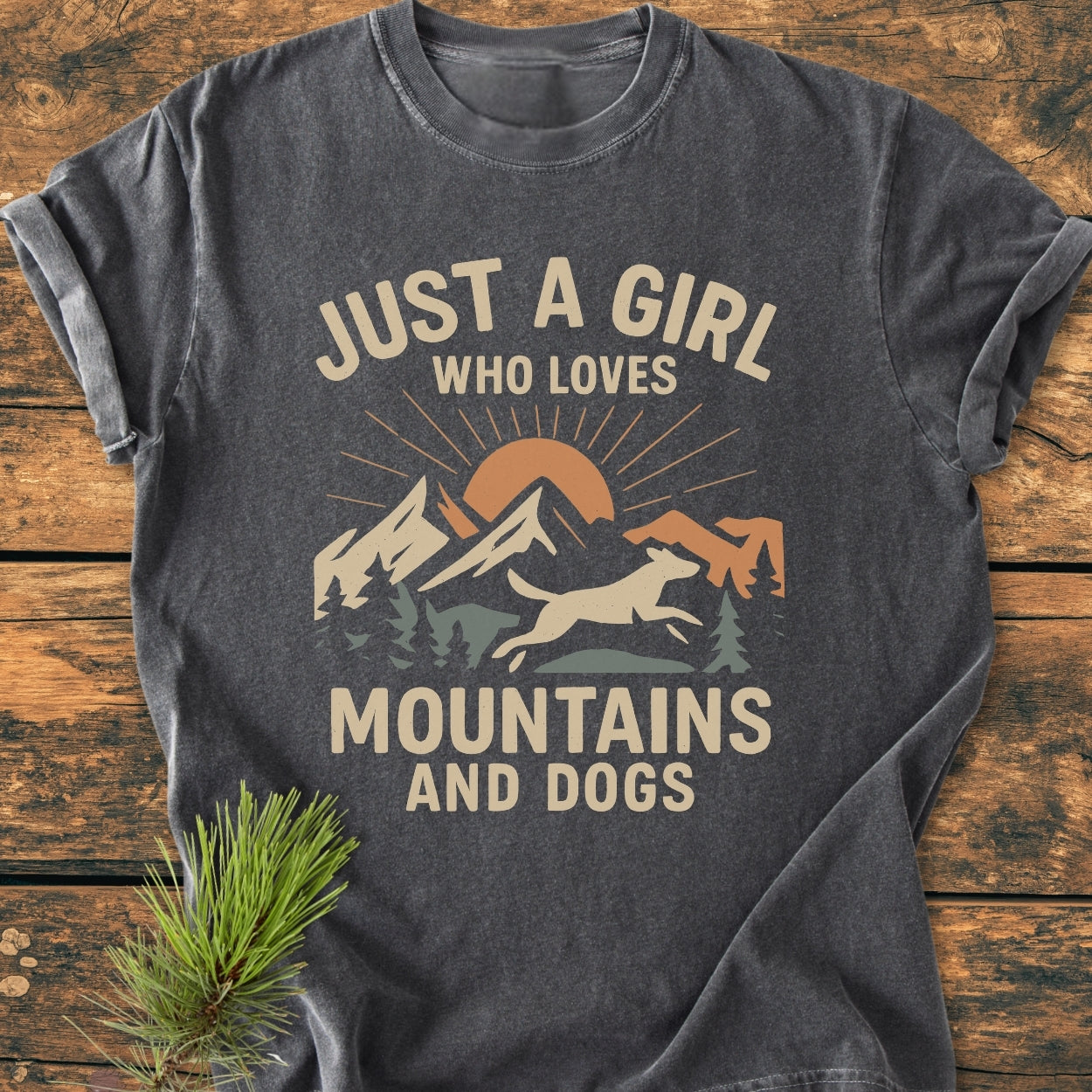 Just A Girl Who Loves Mountains And Dogs - Vintage Wash Tee featuring a stylized sun, mountains, and dog graphic, laid flat on a wooden surface with a pine sprig beside it.