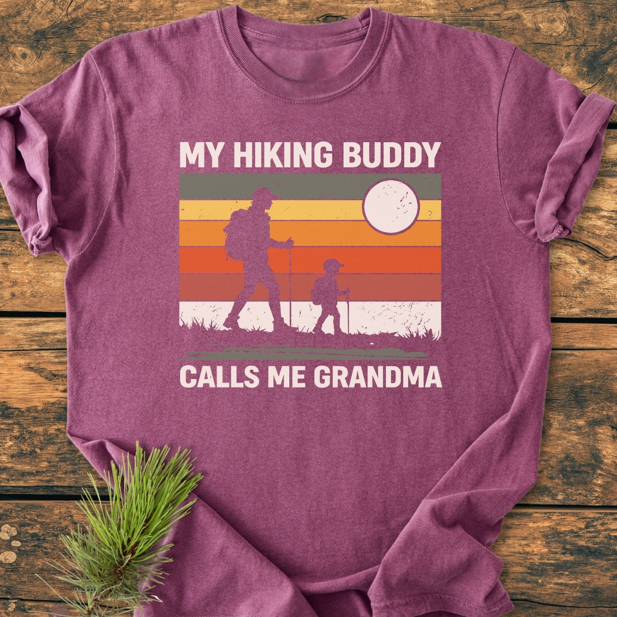 My Hiking Buddy Calls Me Grandma - Vintage Wash Tee features a vintage-style graphic with rainbow stripes, hiking silhouettes, and sun/moon design, laid on wood with pine needles.