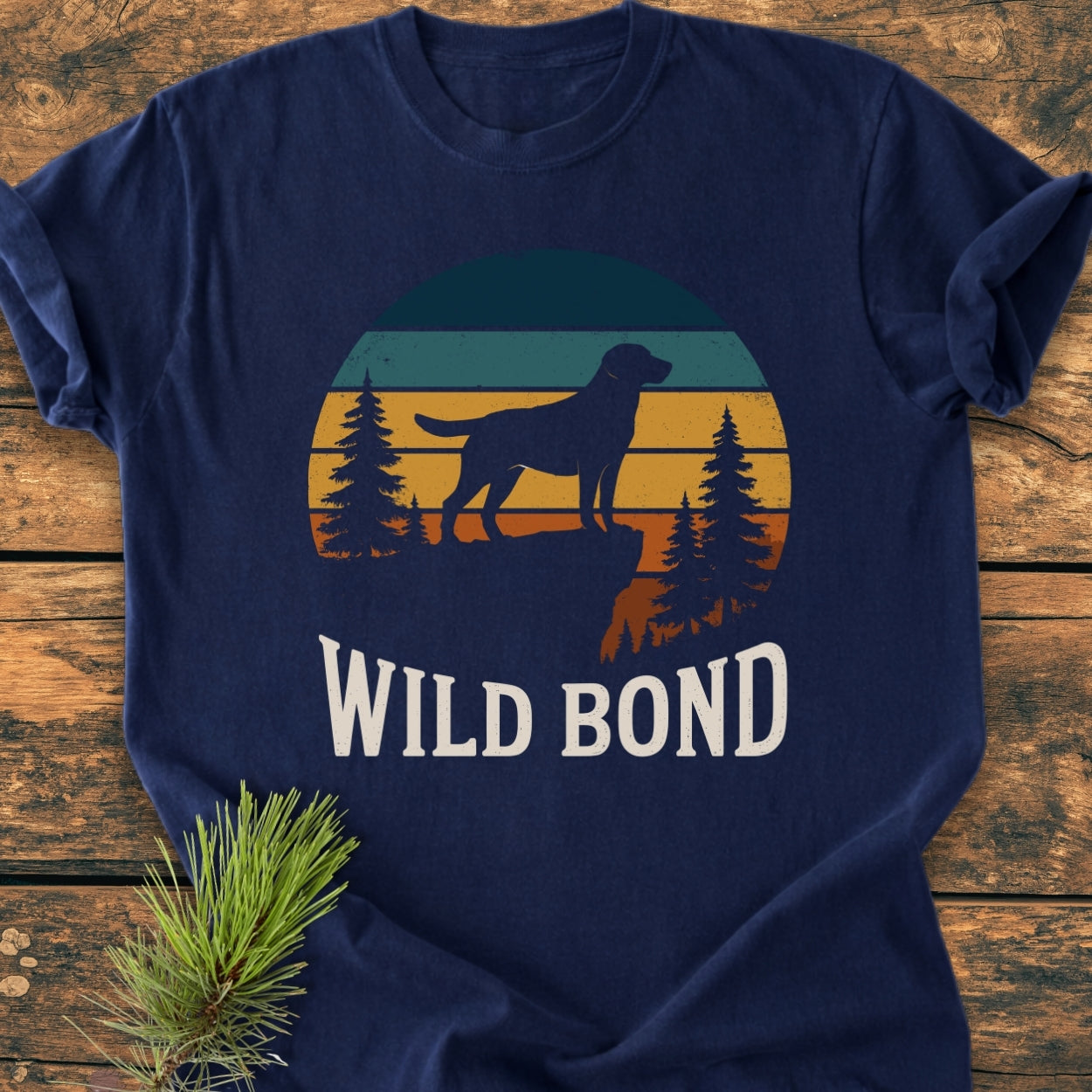 Wild Bond - Vintage Wash Tee: Navy-blue T-shirt with a retro dog graphic on a cliff, surrounded by pine trees. Features WILD BOND text in distressed white font, laid on a wooden surface.