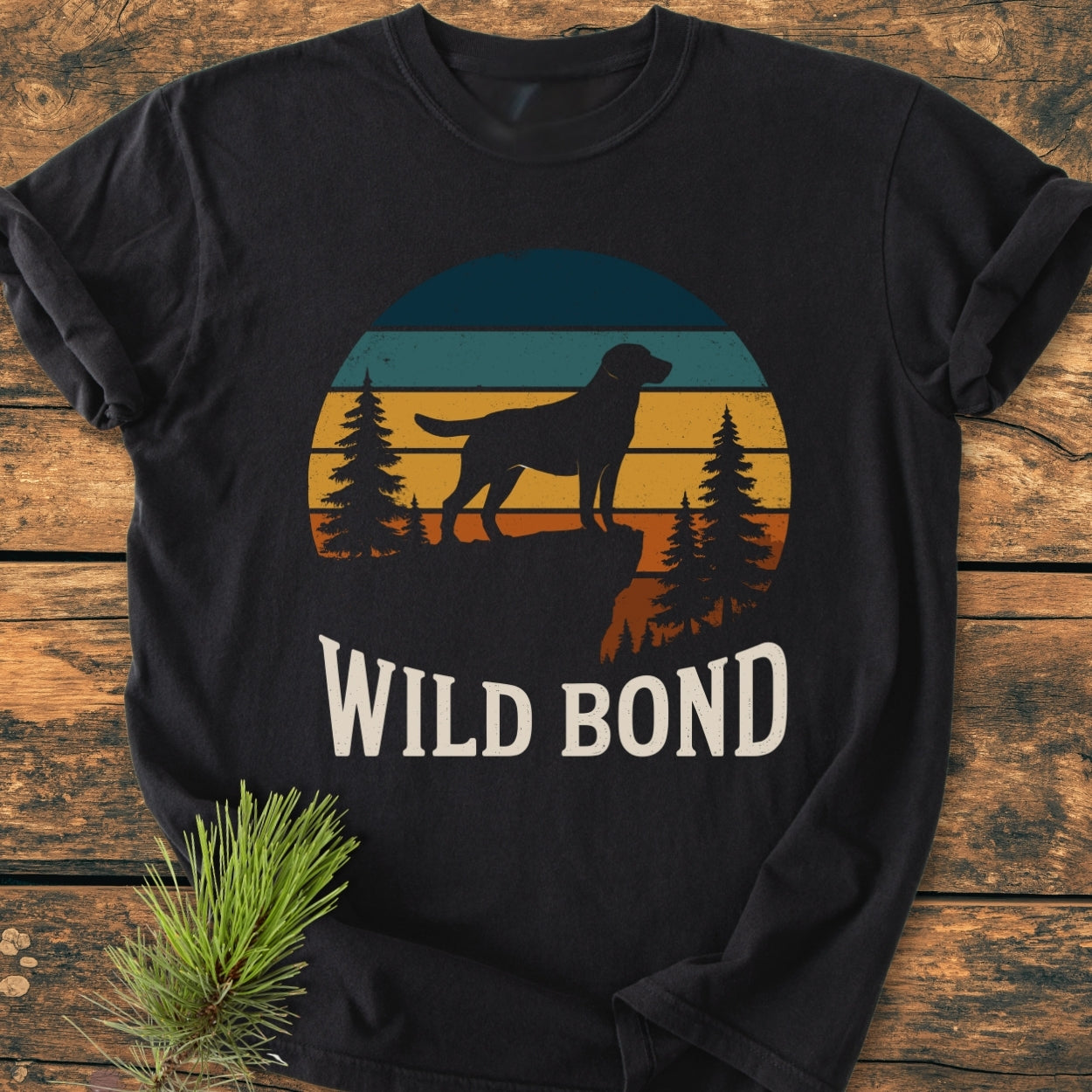 Wild Bond - Vintage Wash Tee featuring a retro sunset with a dog silhouette on a cliff among pine trees, laid on wood, alongside a pine sprig, highlighting its cozy, relaxed fit design.