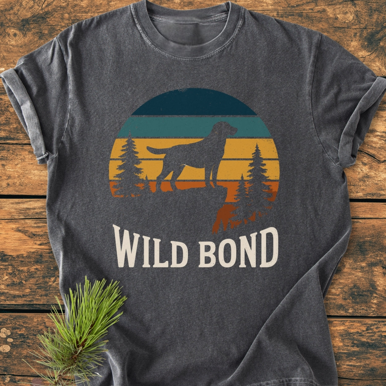 Wild Bond - Vintage Wash Tee features a vintage sunset graphic with a dog and pines, on a charcoal-gray, soft-washed T-shirt made of 100% ring-spun cotton, laid on a wooden surface.