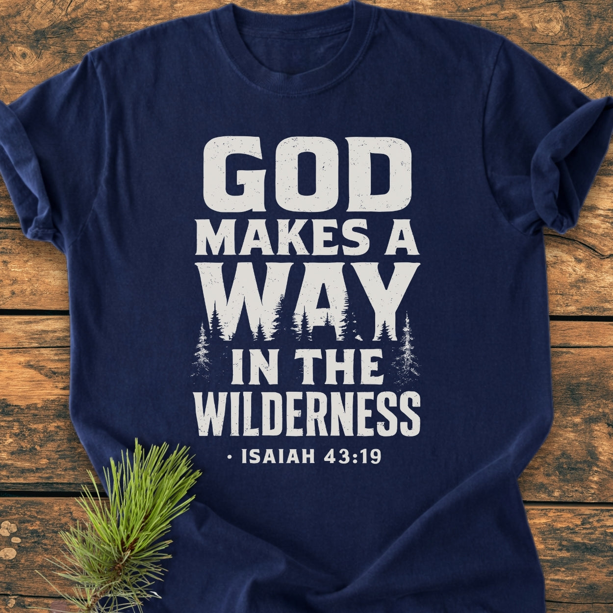Navy-blue God Makes A Way - Vintage Wash Tee featuring bold distressed white lettering with pine tree silhouettes, laid on a wooden surface, showcasing a small pine branch beside it.