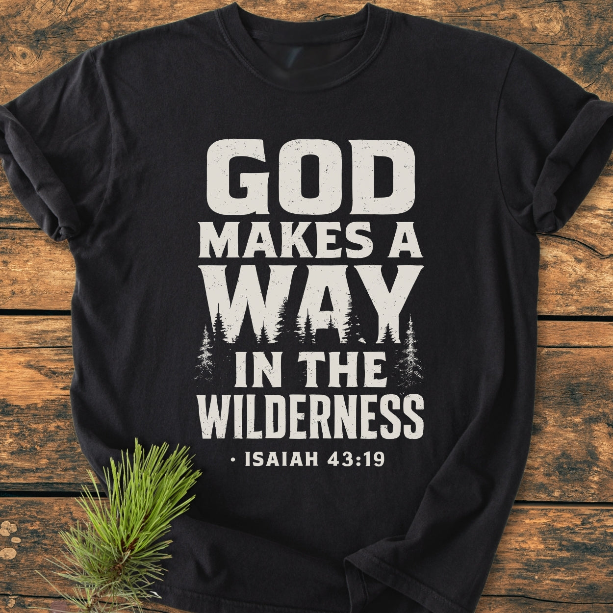 God Makes A Way - Vintage Wash Tee features a bold white print, 'GOD MAKES A WAY IN THE WILDERNESS' with pine tree silhouette, laid on a wooden surface with pine sprig accent.