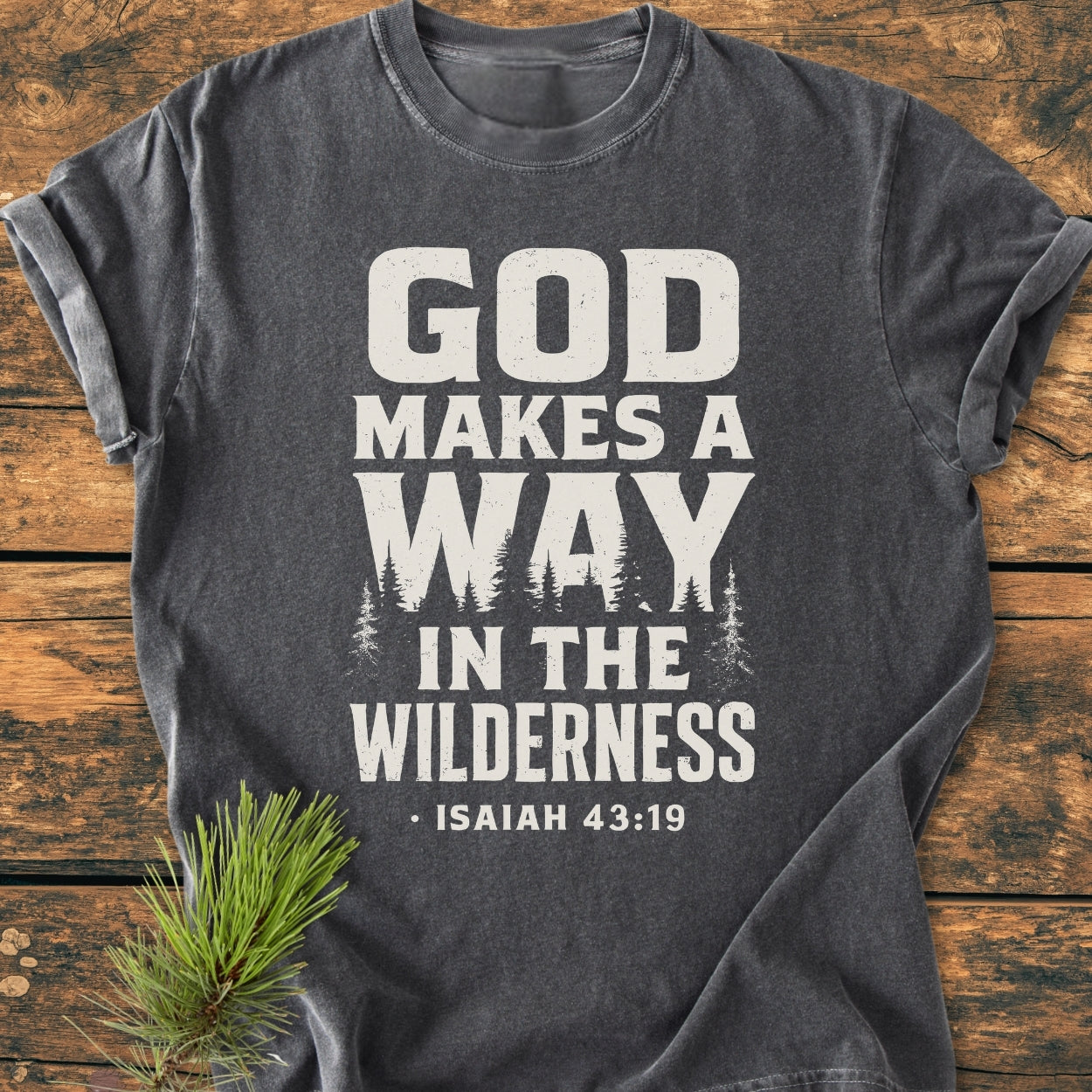 The God Makes A Way - Vintage Wash Tee features distressed white lettering with tree silhouettes, laid on a wooden surface with a pine sprig, emphasizing comfort with its soft, relaxed fit.