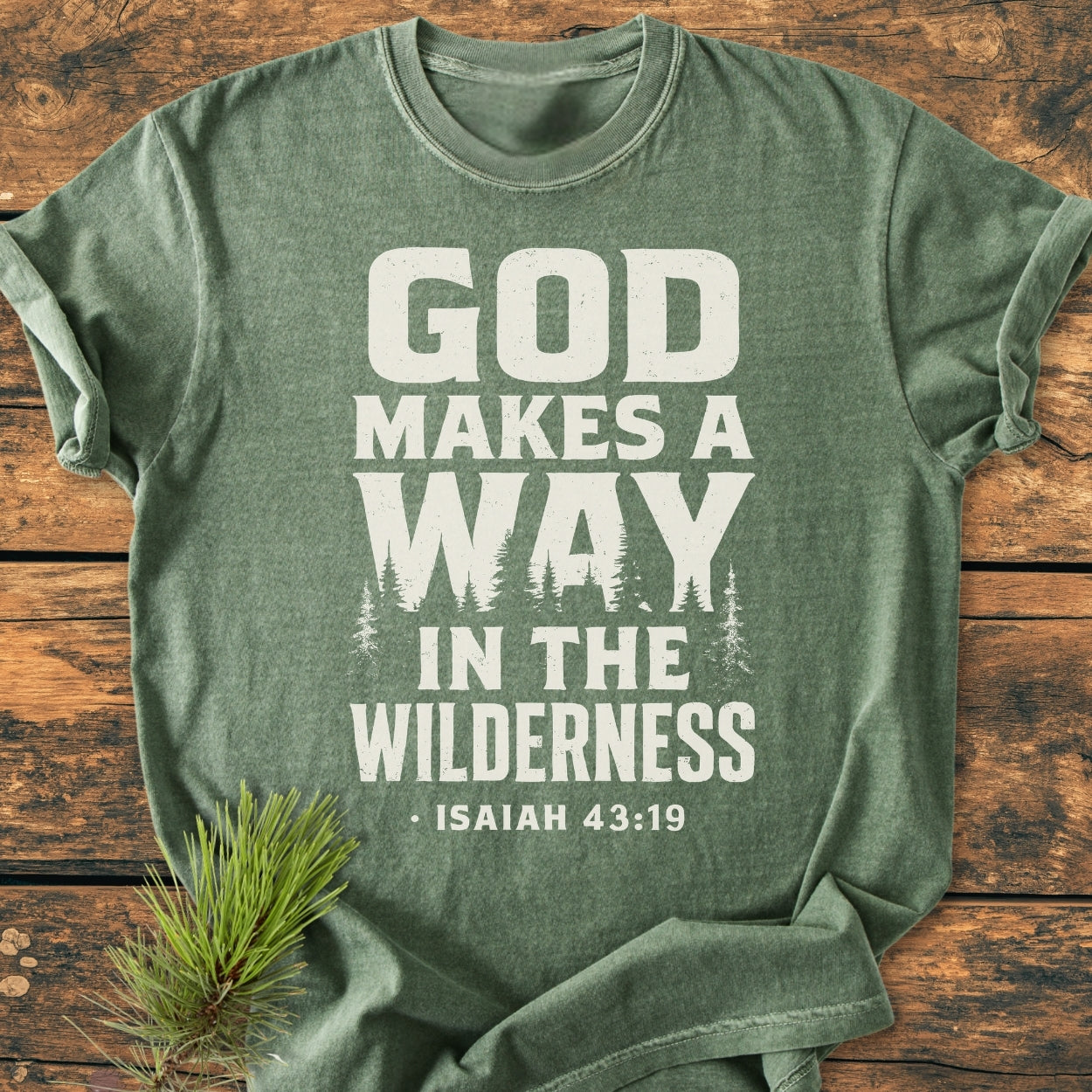 God Makes A Way - Vintage Wash Tee laid flat, featuring a religious verse in bold white letters with pine tree silhouettes, accompanied by a pine sprig on a rustic wooden surface.