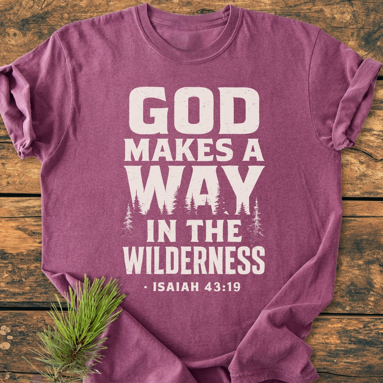 God Makes A Way - Vintage Wash Tee: Folded, plum-colored T-shirt on wood, featuring distressed white text and pine tree silhouettes. A pine branch accentuates the tee's rustic, comforting aesthetic.