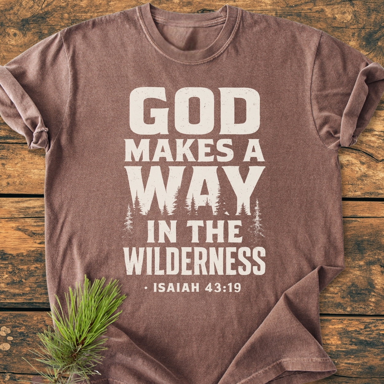 God Makes A Way - Vintage Wash Tee featuring off-white lettering with tree silhouette on a brown heathered fabric, laid flat with a pine bough accentuating the wilderness theme.