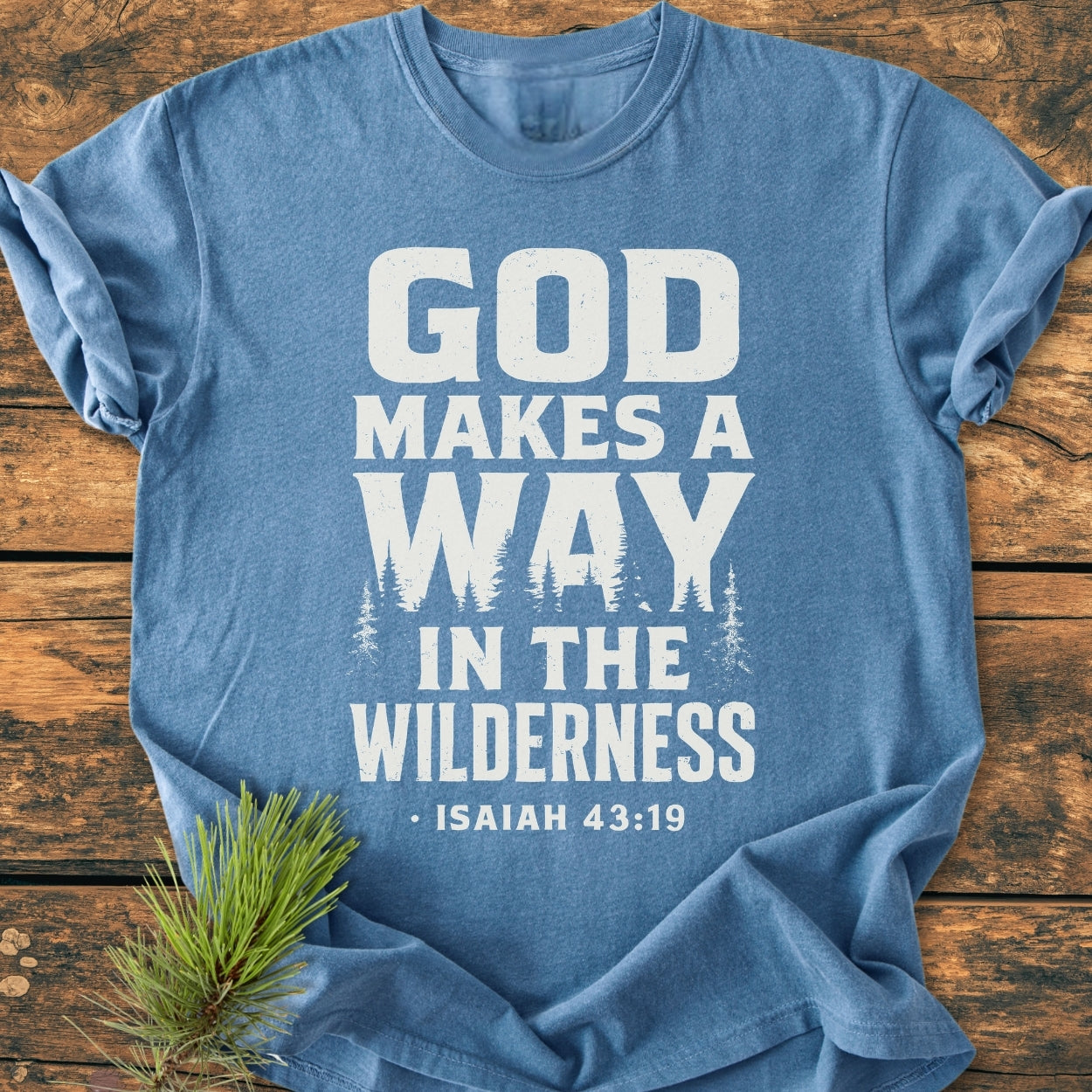 God Makes A Way - Vintage Wash Tee features bold white text and tree silhouettes on a heather-blue background, laid on wood. Accompanied by a small pine sprig for a rustic touch.