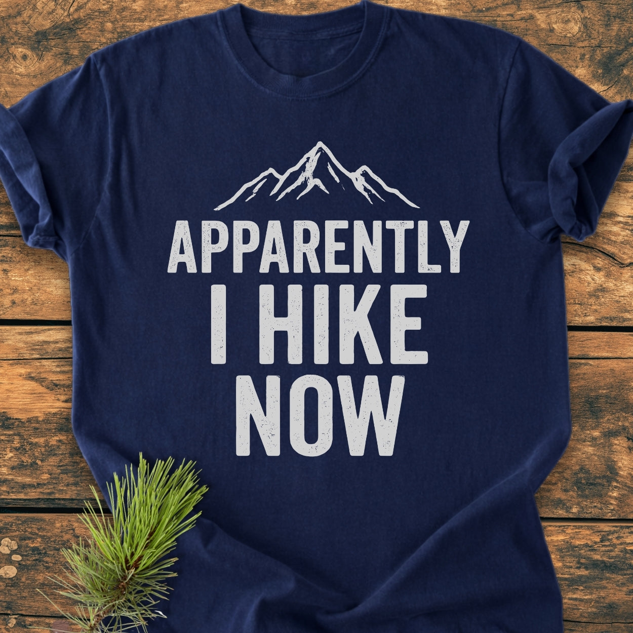 Navy-blue Apparently Hiking - Vintage Wash Tee features a white mountain peak design and bold text. Resting on a wooden surface with a pine branch accentuating the garment's rustic appeal.