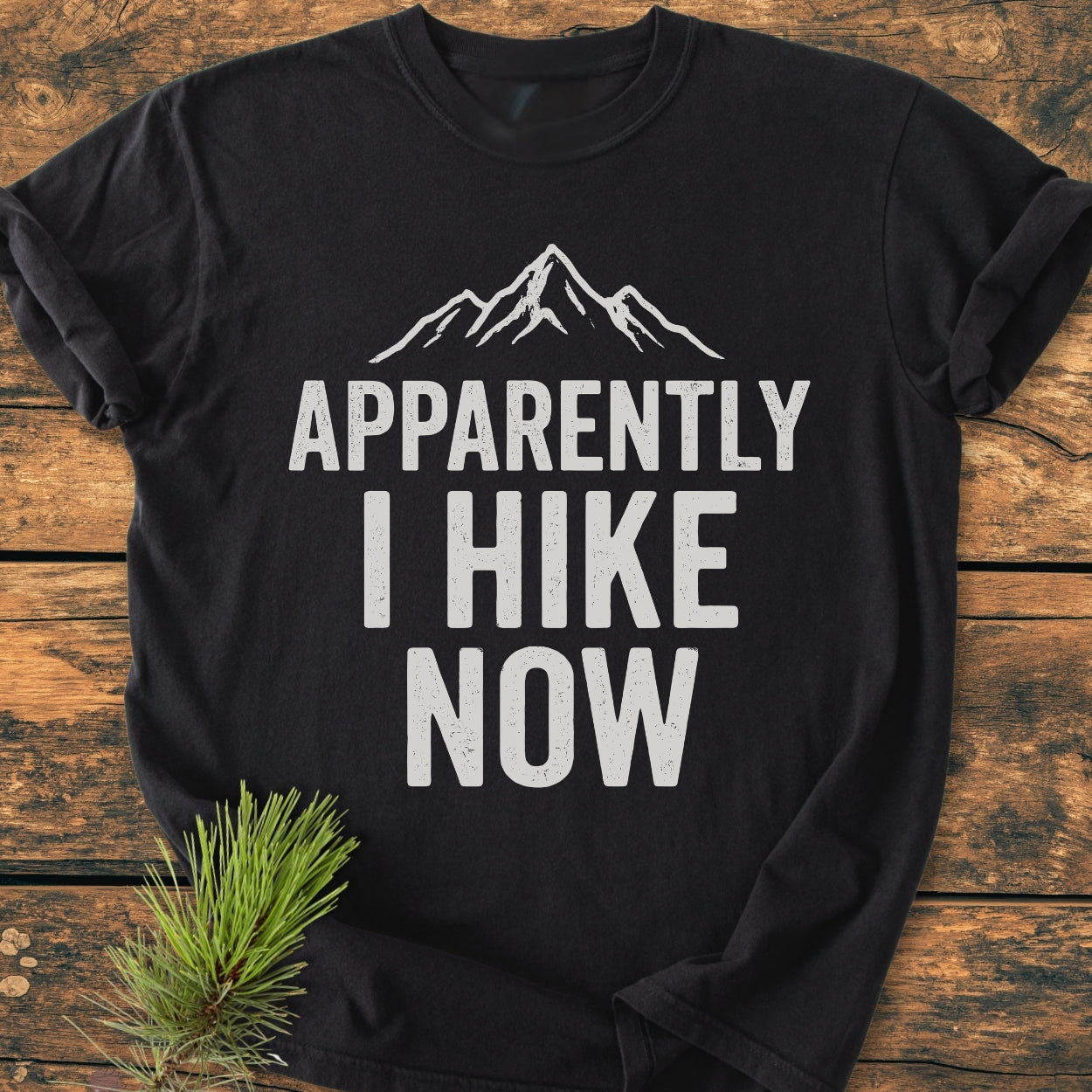 Apparently Hiking - Vintage Wash Tee on wooden surface, featuring a mountain-peak graphic and bold text. A pine sprig accents the soft, relaxed-fit, black T-shirt made of durable, pre-shrunk ring-spun cotton.