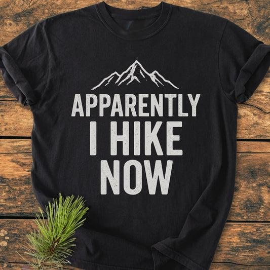 Apparently Hiking - Vintage Wash Tee on wooden surface, featuring a mountain-peak graphic and bold text. A pine sprig accents the soft, relaxed-fit, black T-shirt made of durable, pre-shrunk ring-spun cotton.