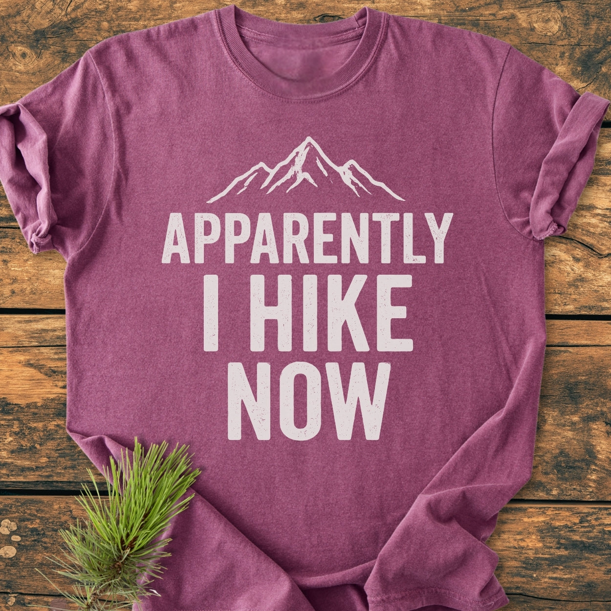 Apparently Hiking - Vintage Wash Tee featuring a white mountain peak graphic and text “APPARENTLY I HIKE NOW” on a wooden surface, accented with a pine sprig for an outdoorsy touch.