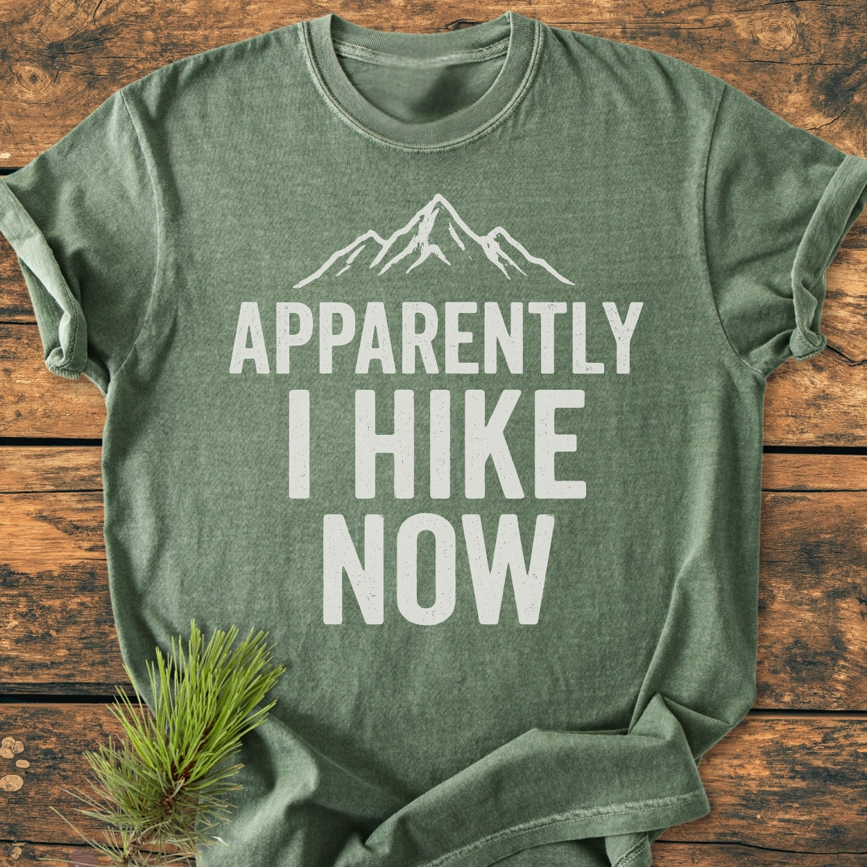 Apparently Hiking - Vintage Wash Tee, green heather-style laid flat, features a white mountain outline and slogan. Accented by a pine sprig, showcasing Comfort Colors' relaxed, durable, 100% cotton design.