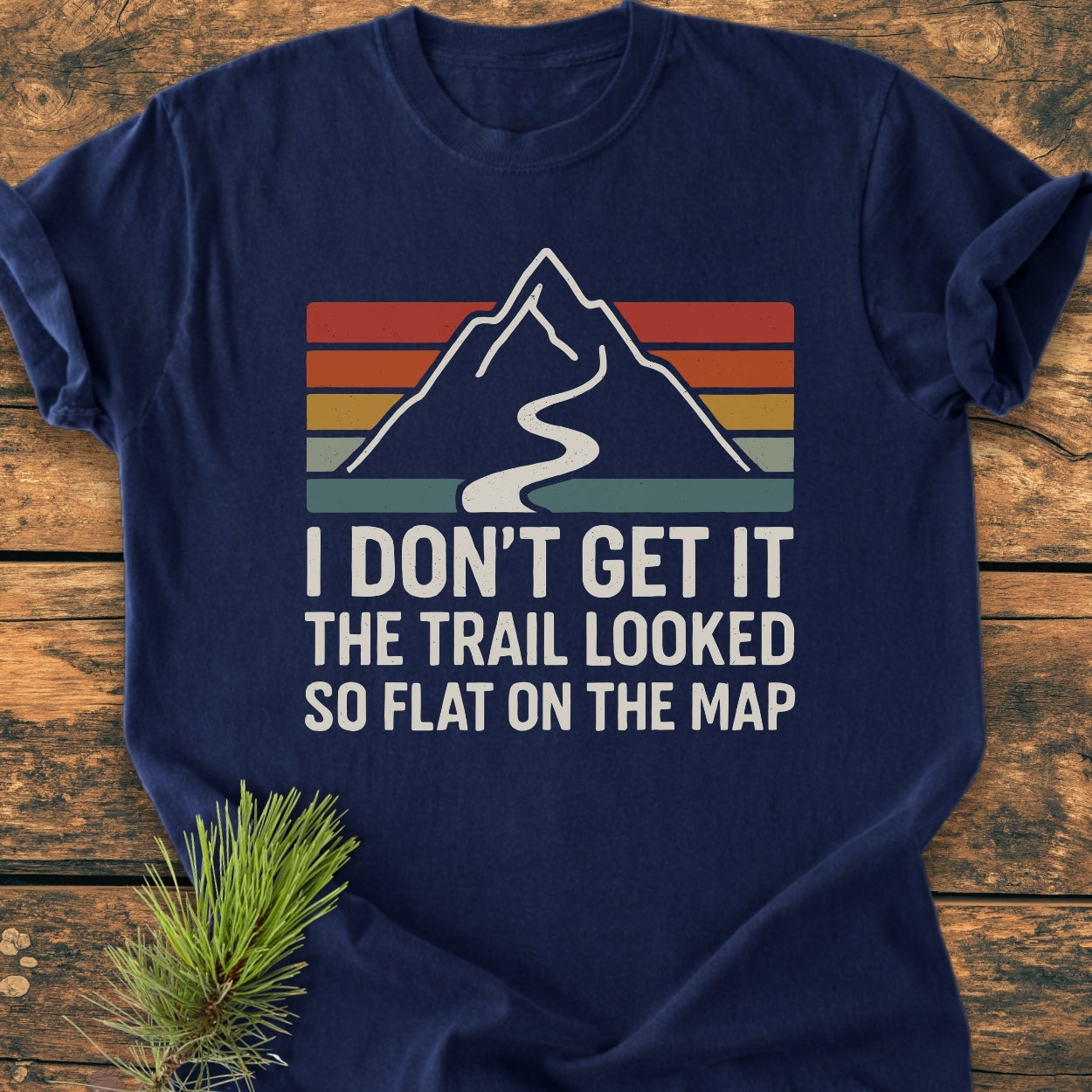 The I Don't Get It - Vintage Wash Tee features a retro mountain graphic with a humorous slogan, printed on a navy-blue, soft-washed T-shirt, lying on a wooden surface with a pine sprig.