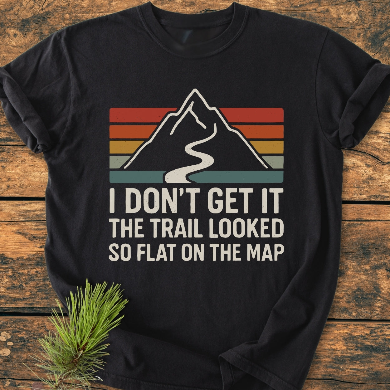 I Don't Get It - Vintage Wash Tee featuring a mountain graphic with retro stripes and a humorous slogan, laid on a wooden surface, beside a small pine sprig.