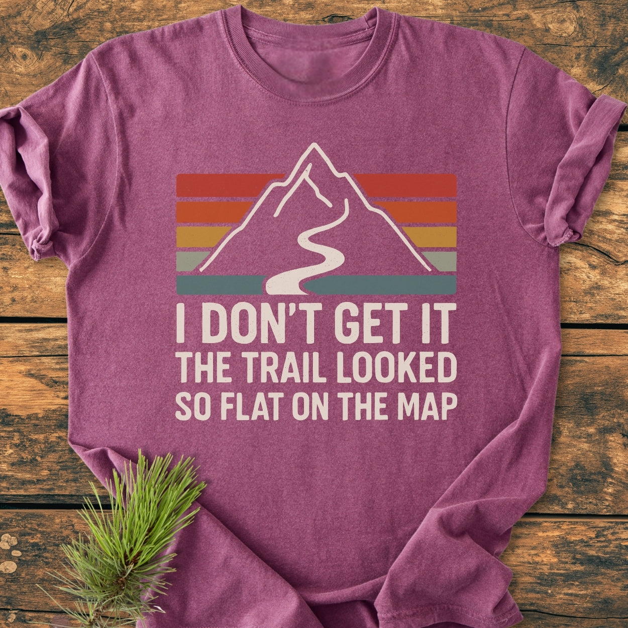 I Don't Get It - Vintage Wash Tee features a mountain graphic on a folded purple shirt, laid on wood with a pine branch. Crafted from soft, ring-spun cotton for comfort and durability.
