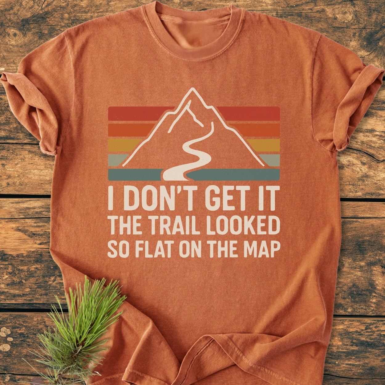 The I Don't Get It - Vintage Wash Tee features a stylized mountain design with humorous text on a rust-orange T-shirt, displayed on a wooden surface with a pine sprig.