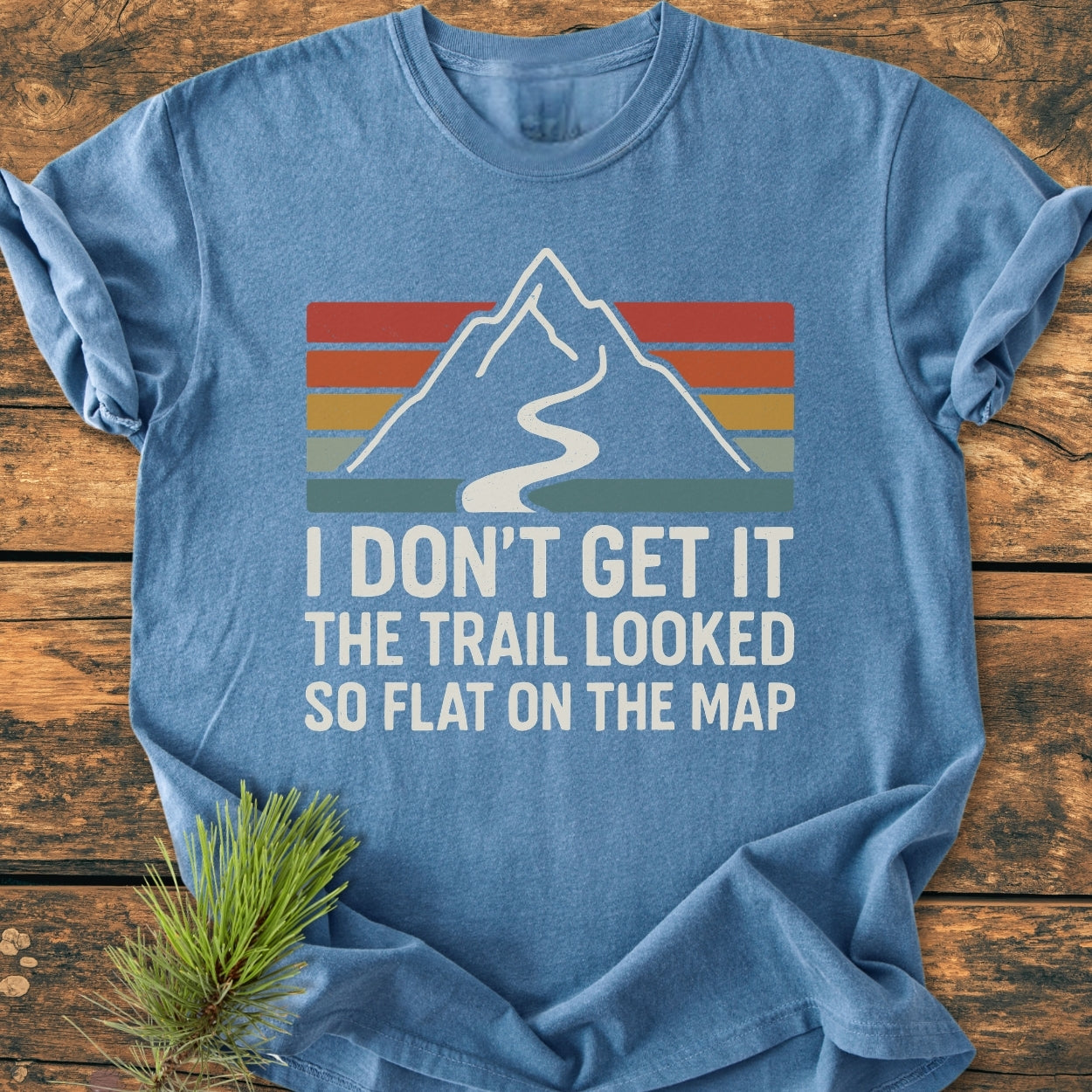 Heather-blue I Don't Get It - Vintage Wash Tee featuring a retro mountain graphic with a winding trail. Includes a humorous slogan and a pine-needle sprig beside it, laid on wood surface.