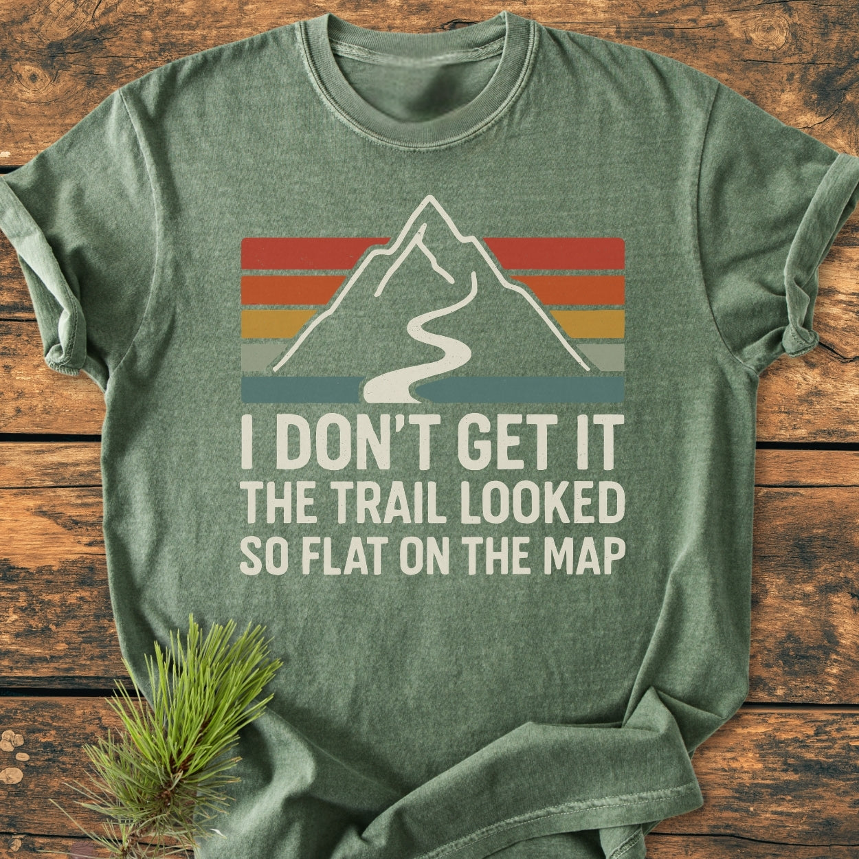 I Don't Get It - Vintage Wash Tee features a mountain graphic with a trail and a humorous slogan, laid on a wooden surface, showcasing its heather-green hue and retro style.
