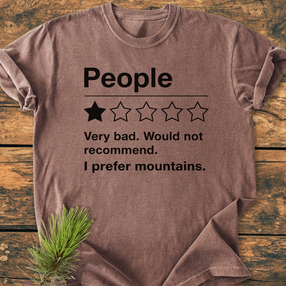 Brown T-shirt titled I Prefer Mountains - Vintage Wash Tee with humorous text and a one-star rating graphic, laid on a wooden surface with a sprig of fir in the corner.