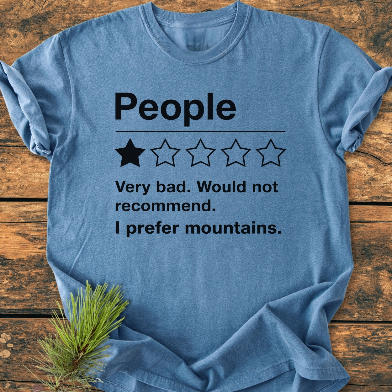 I Prefer Mountains - Vintage Wash Tee: Heather-blue T-shirt with mock negative review, laid on rustic wood. Pine sprig accentuates nature theme, emphasizing comfort and relaxed fit of the garment-dyed cotton shirt.