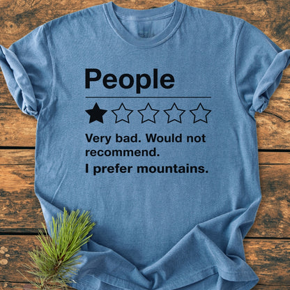 I Prefer Mountains - Vintage Wash Tee: Heather-blue T-shirt with mock negative review, laid on rustic wood. Pine sprig accentuates nature theme, emphasizing comfort and relaxed fit of the garment-dyed cotton shirt.