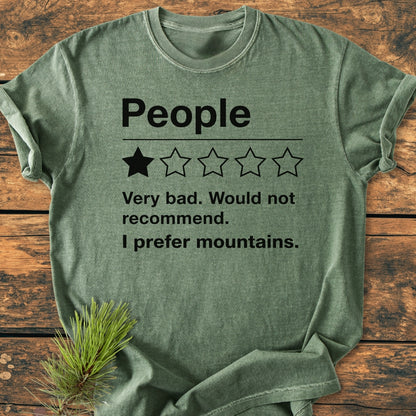 I Prefer Mountains - Vintage Wash Tee: Heather-green T-shirt with humorous mock review, emphasizing mountain preference. Laid flat on wood, complemented by a pine sprig, exuding an outdoorsy vibe.