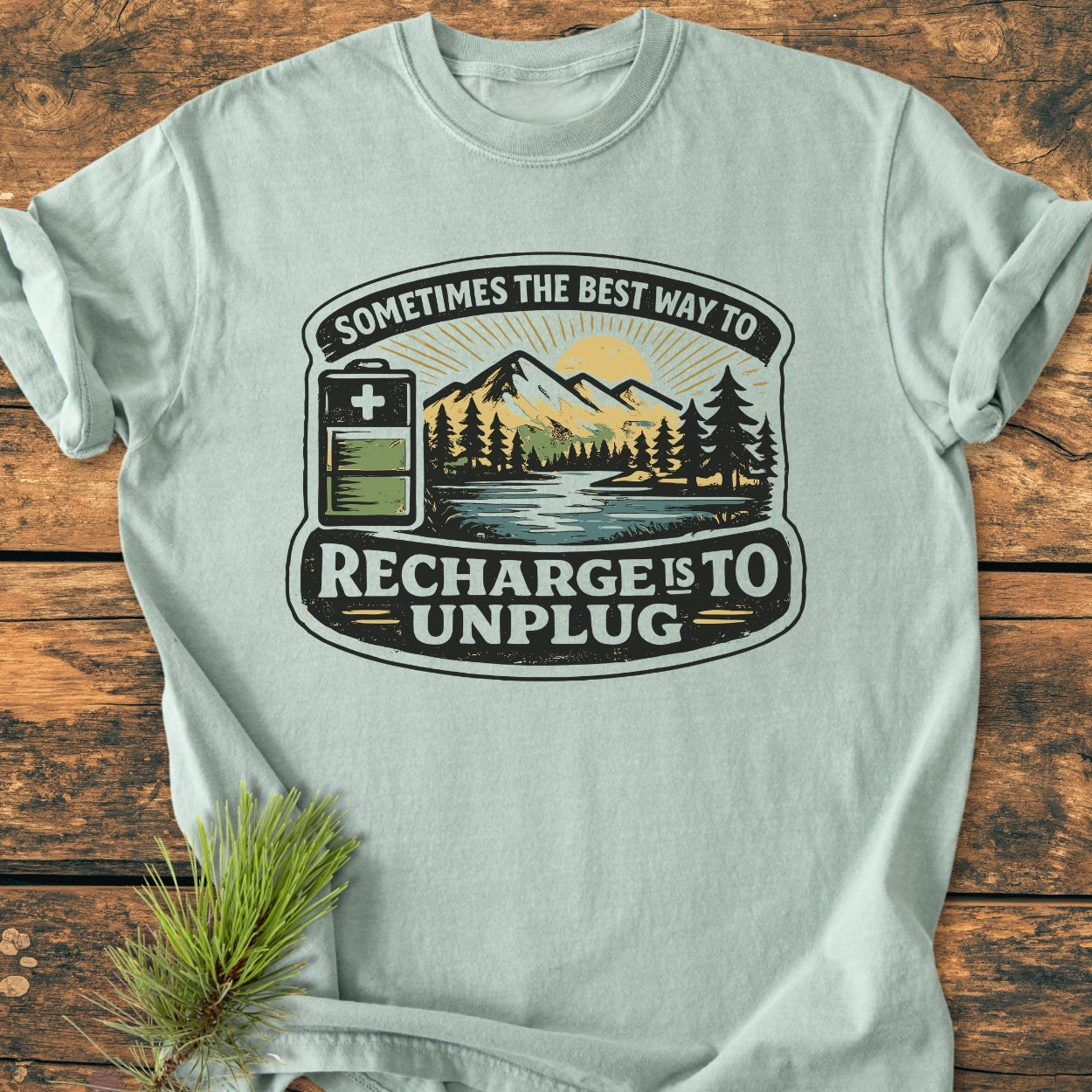 Unplug To Recharge - Vintage Wash Tee features a stylized nature scene with a slogan, laid flat on wood, alongside an evergreen sprig, showcasing its eco-friendly theme and relaxed fit.