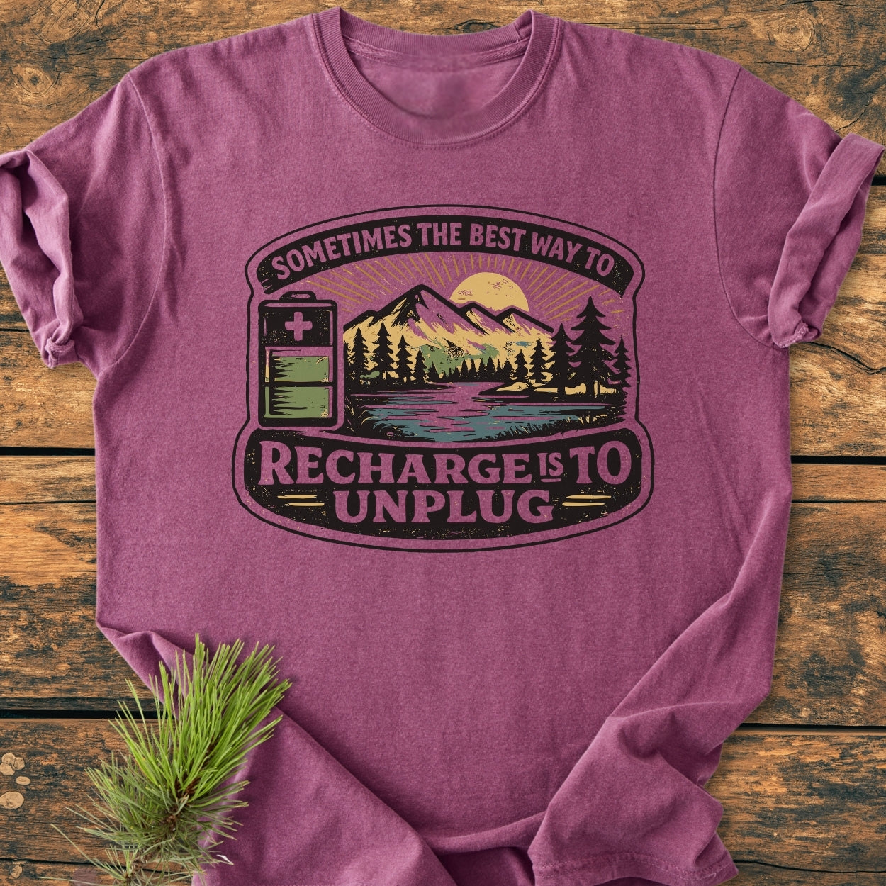Unplug To Recharge - Vintage Wash Tee: A folded purple-mauve T-shirt on wood, featuring a retro graphic with a battery icon, mountain range, and slogan emphasizing the importance of recharging by unplugging.